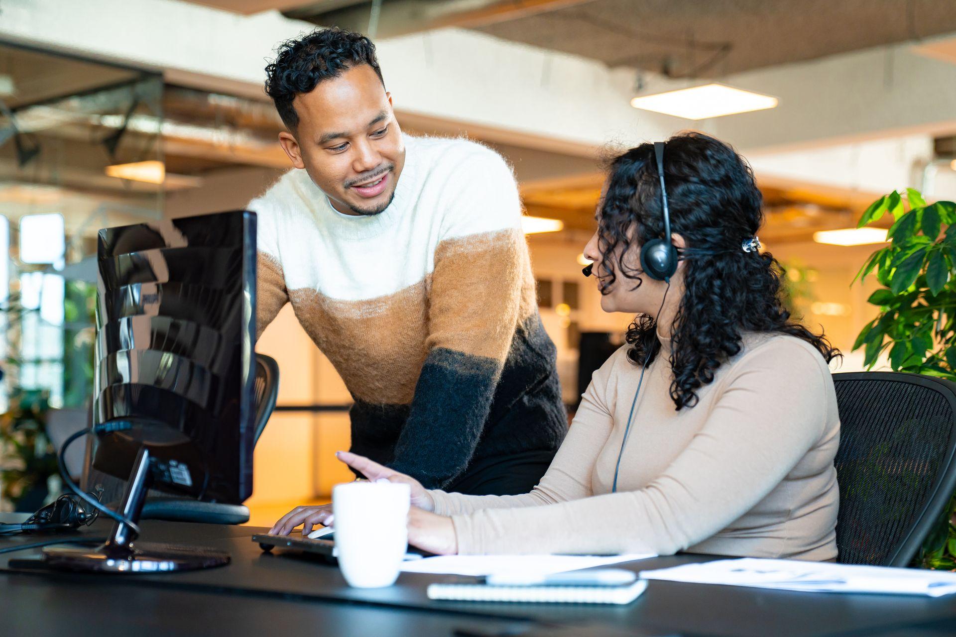 Two colleagues discussing at a desk, one wearing a headset, in a modern office setting.